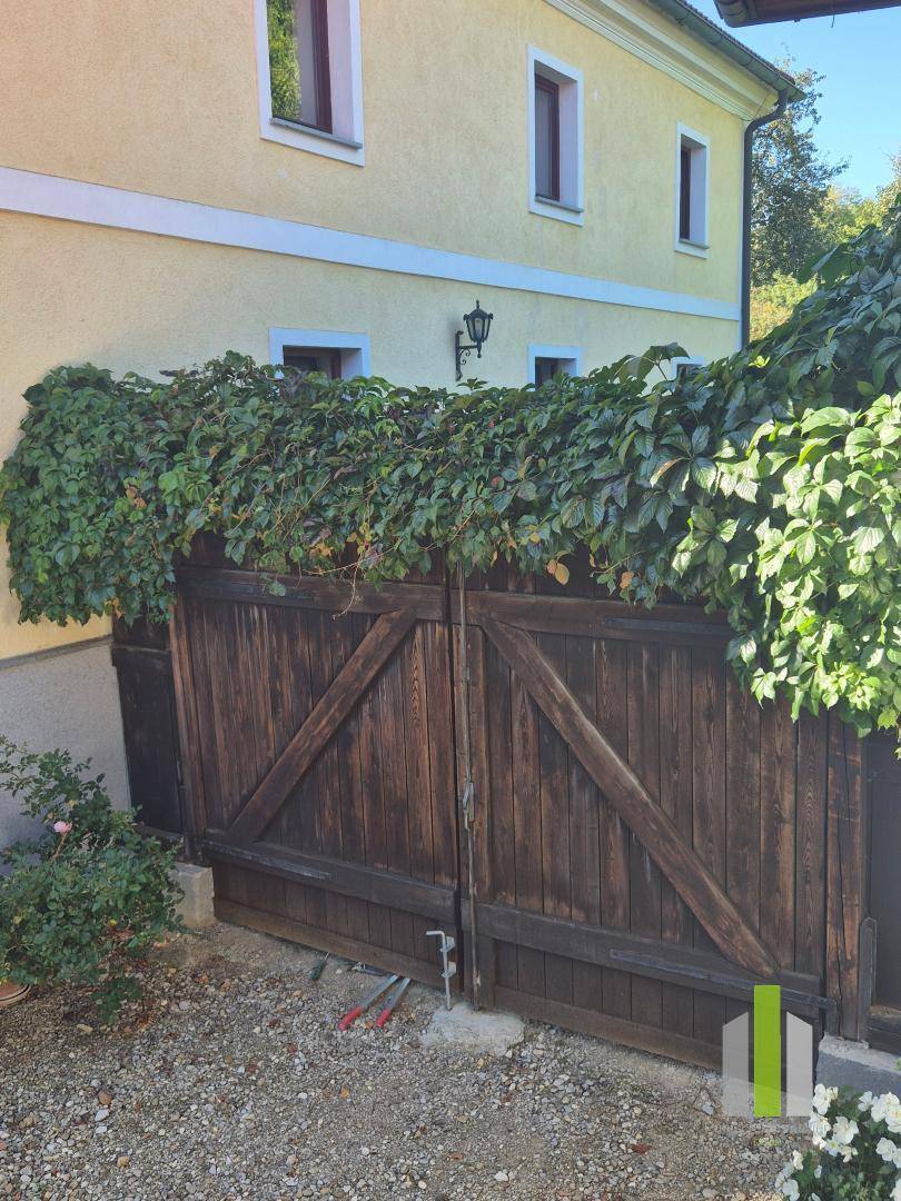 Rustikales Holztor, teilweise mit Efeu bewachsen, neben der gelben Hausfassade. 