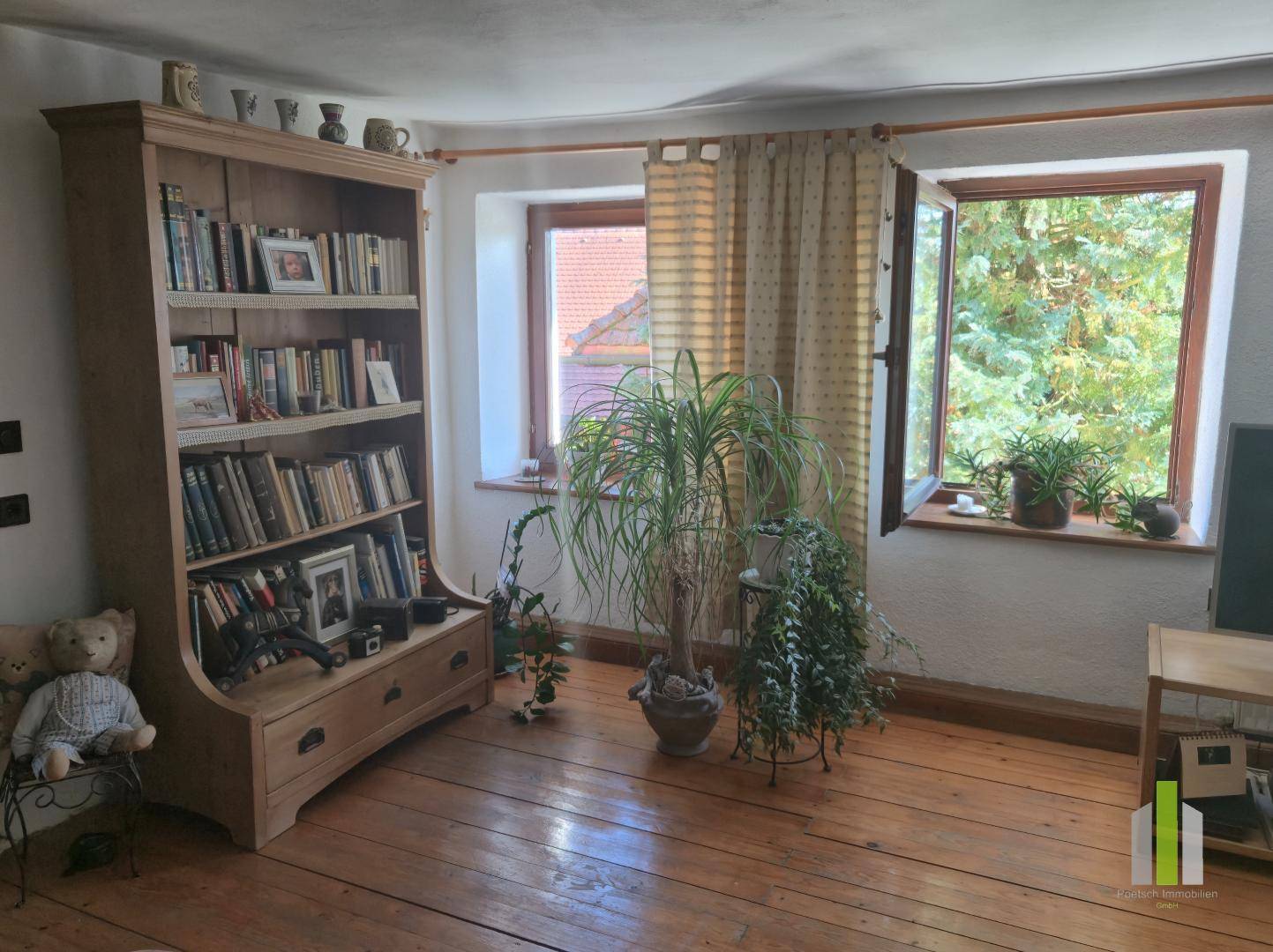 Heller Raum mit Holzdielenboden, Bücherregal und Pflanzen am Fenster mit Blick ins Grüne.