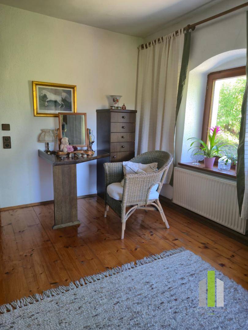 Helles Zimmer mit Holzdielenboden, Korbsessel und einem kleinen Schreibtisch am Fenster.