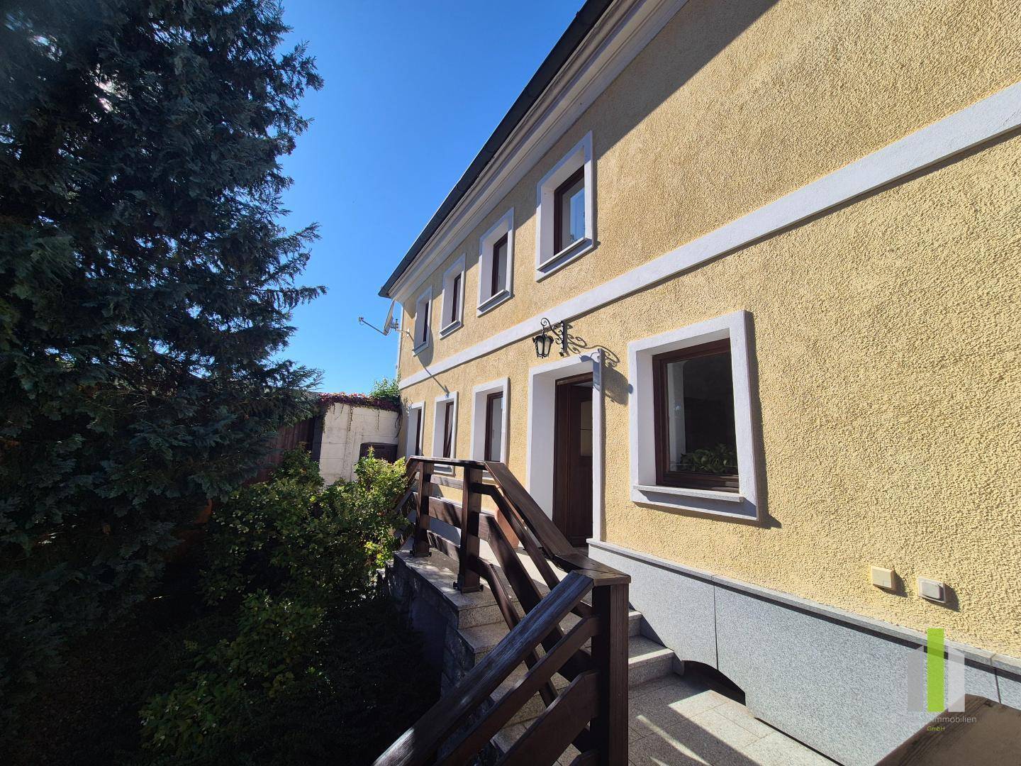 Seitlicher Eingang des Hauses mit Holztreppe und Geländer unter blauem Himmel. 