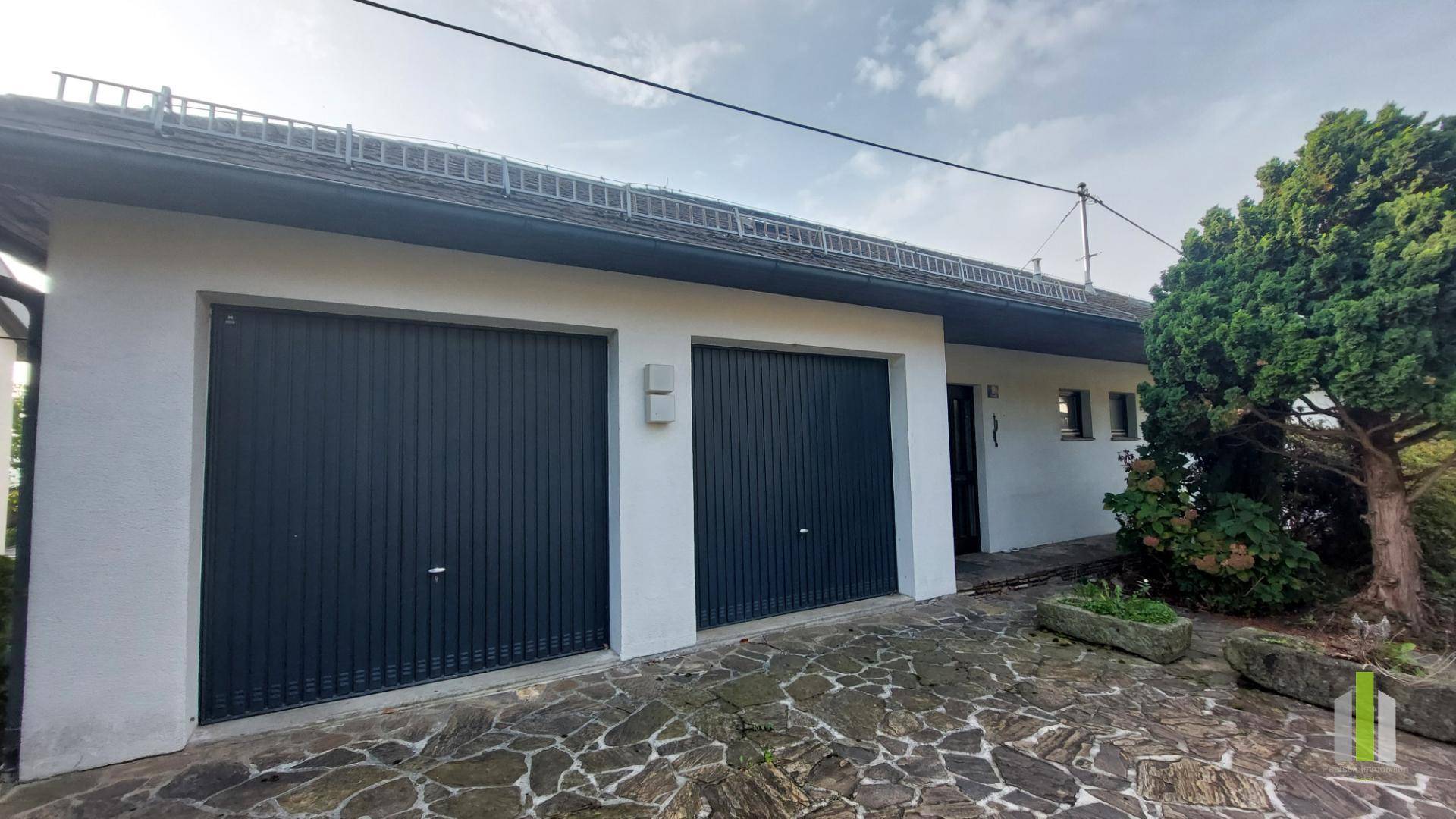 Vorderansicht des Hauses mit zwei dunklen Garagentoren und gepflastertem Vorplatz.