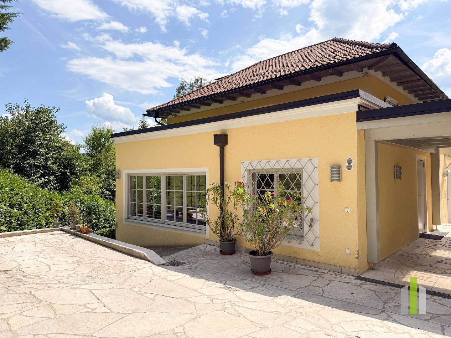 Gelbe Hausfassade mit gepflasterter Terrasse und üppiger grüner Bepflanzung.