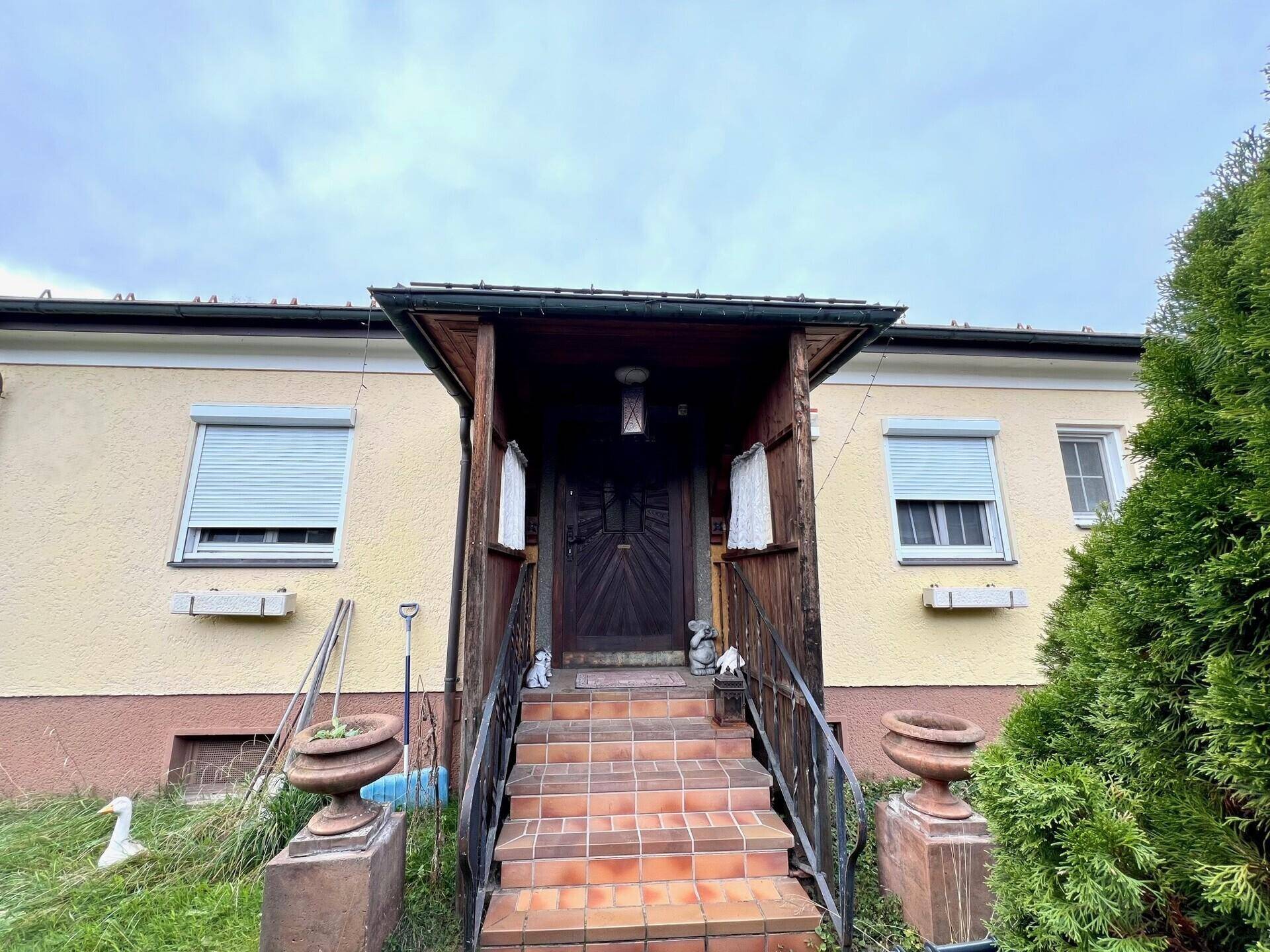 Haupteingang des Hauses mit überdachtem Vorbau, gefliesten Stufen und Fensterläden an den Fenstern.