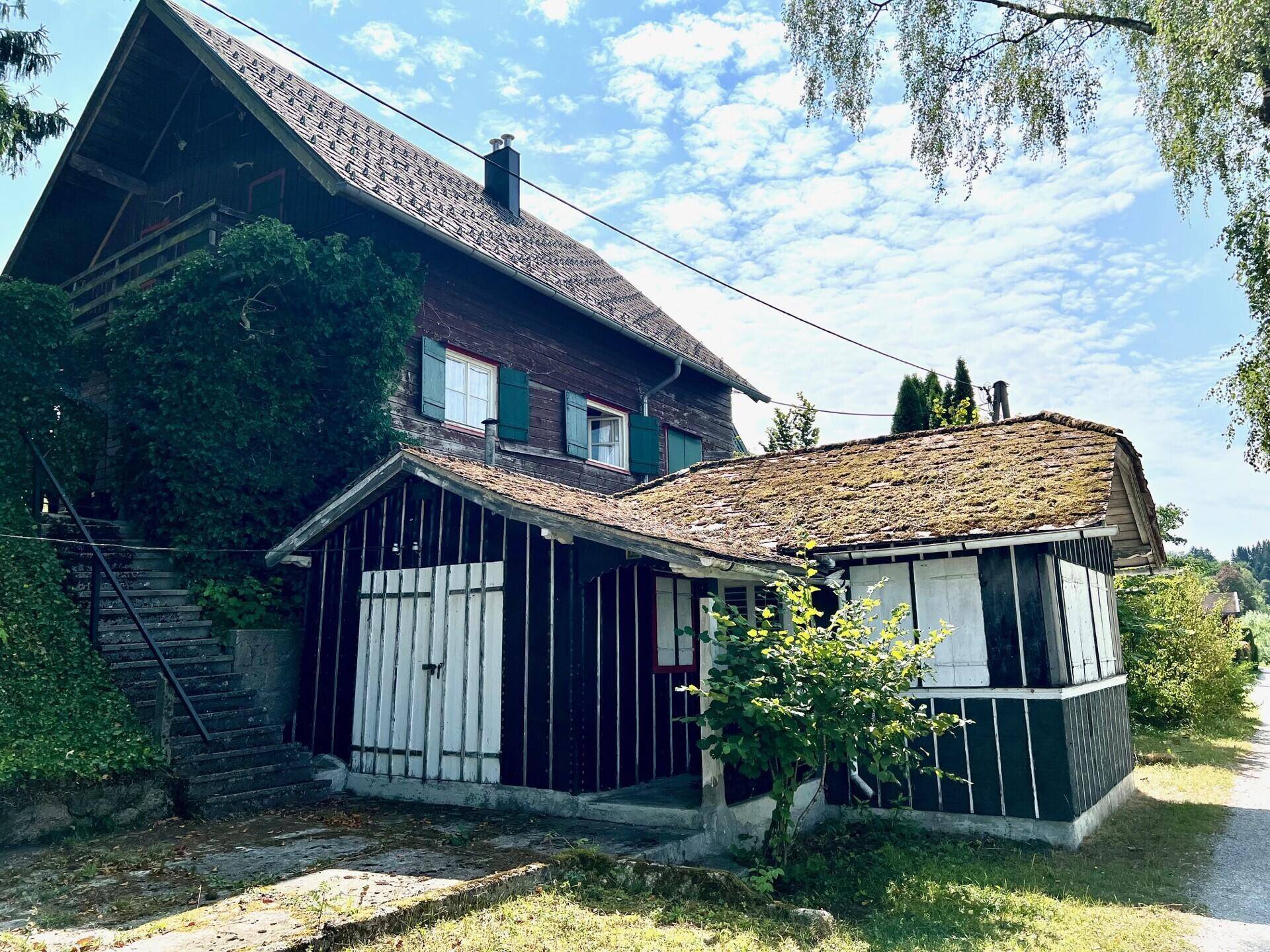 Rustikales Holzhaus mit einem Anbau und moosbewachsenem Dach, umgeben von üppigem Grün und Bäumen.