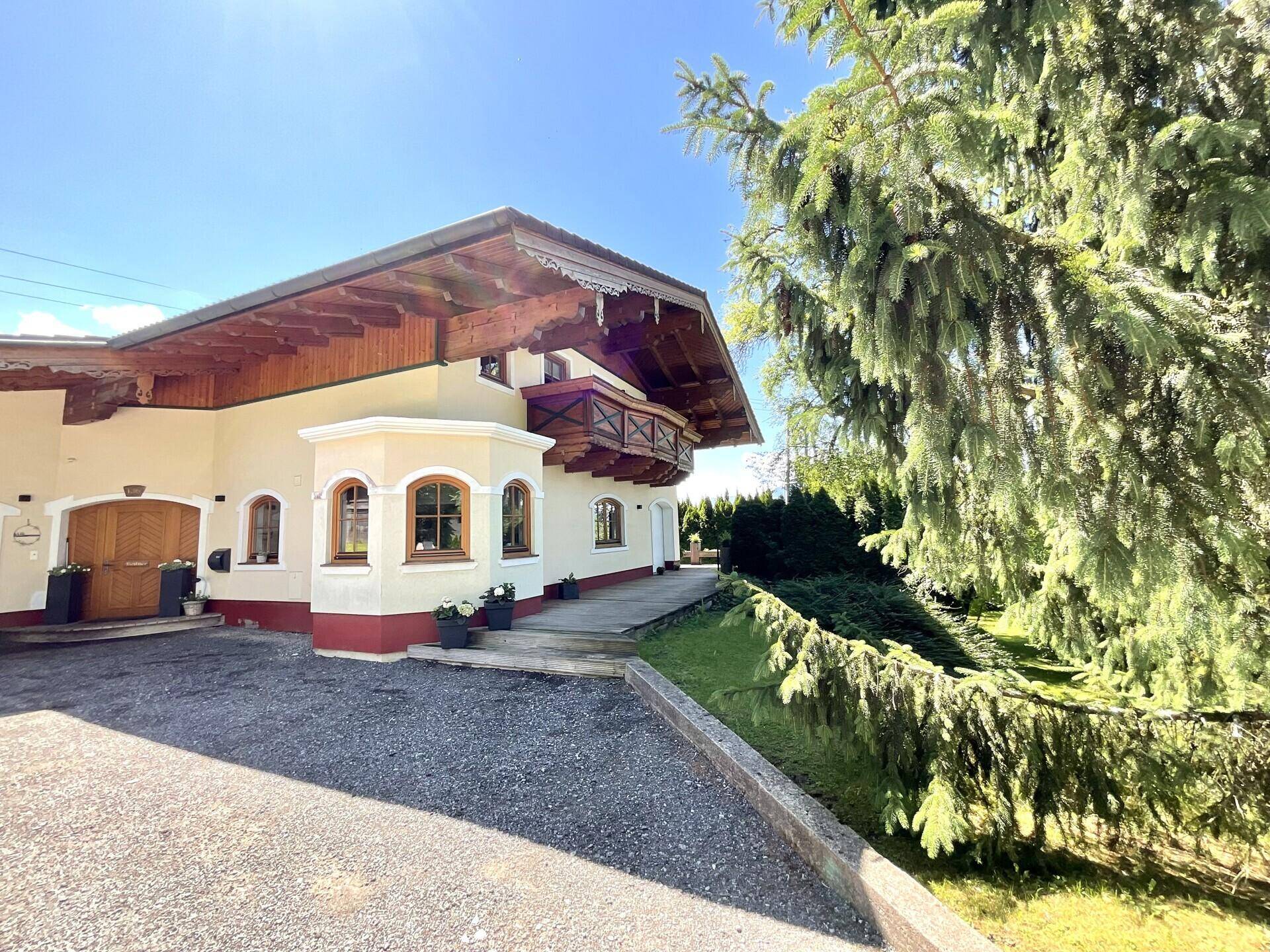 Traditionelles Haus mit Holzbalkon und Kiesauffahrt, umgeben von hohen Bäumen unter blauem Himmel.