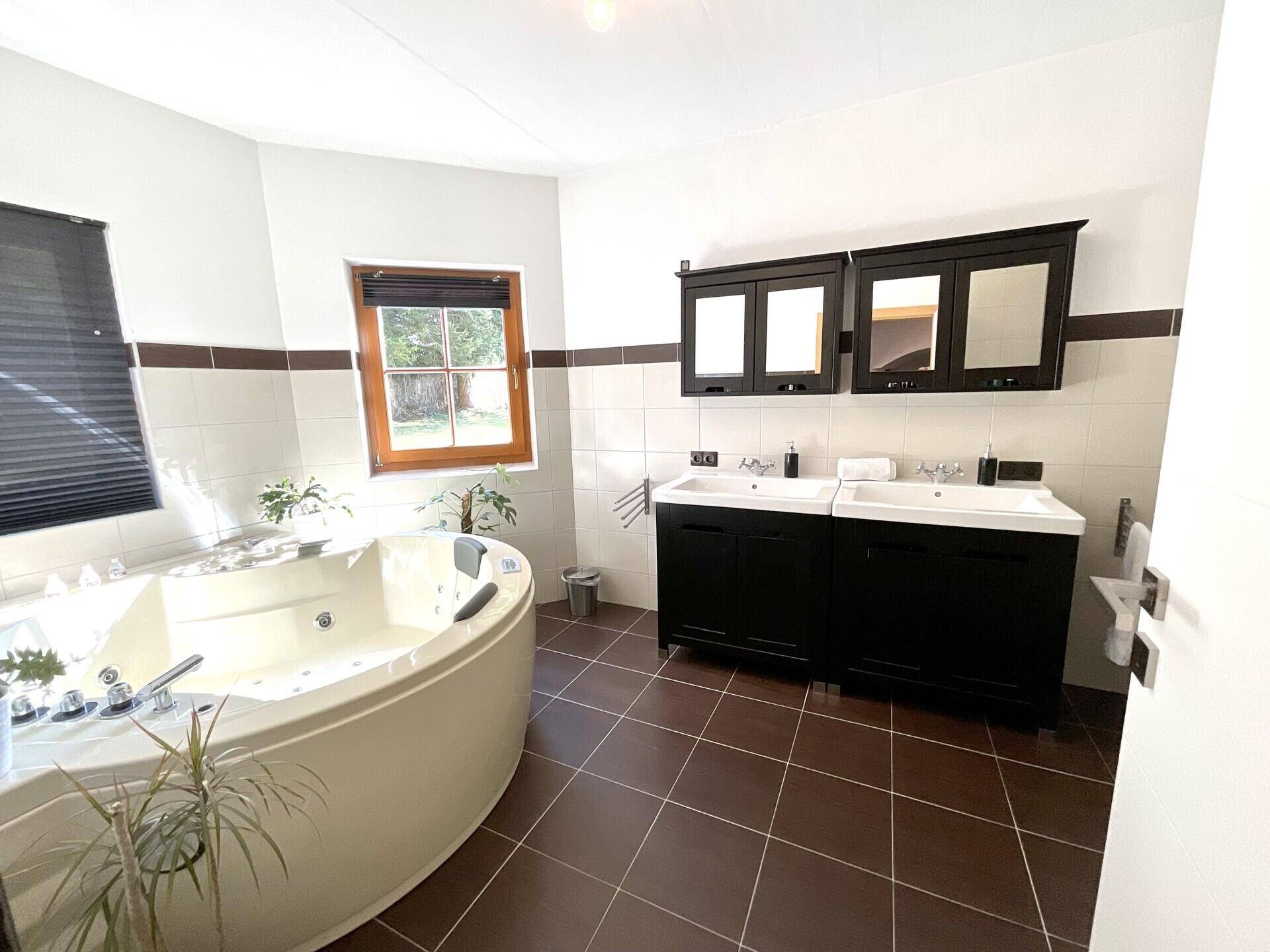 Geräumiges Badezimmer mit Whirlpool-Badewanne, Doppelwaschbecken und braunen Bodenfliesen sowie einem Fenster.