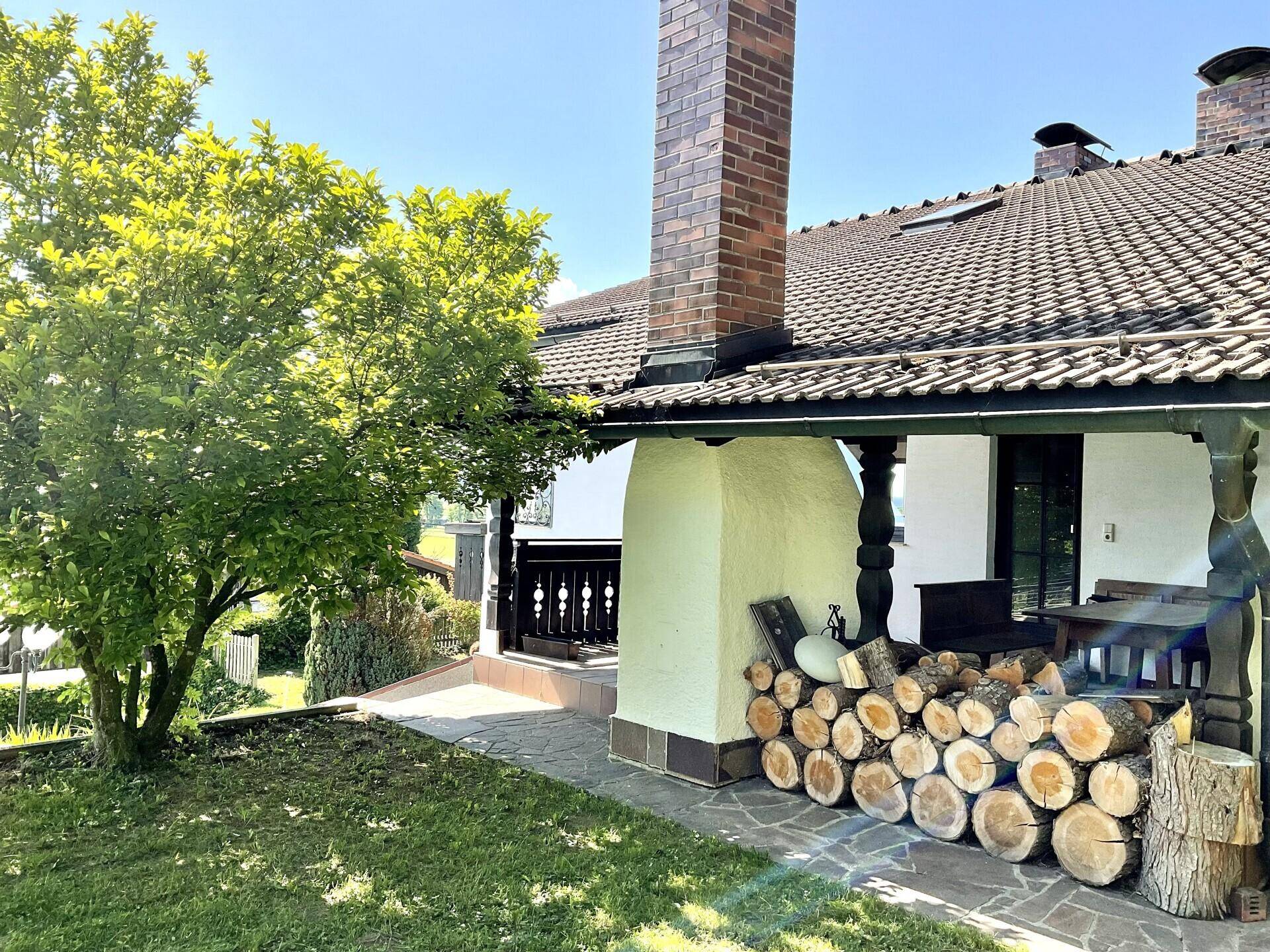Seitlicher Außenbereich des Hauses mit Holzstapel und traditionellem Kamin unter einem Vordach.