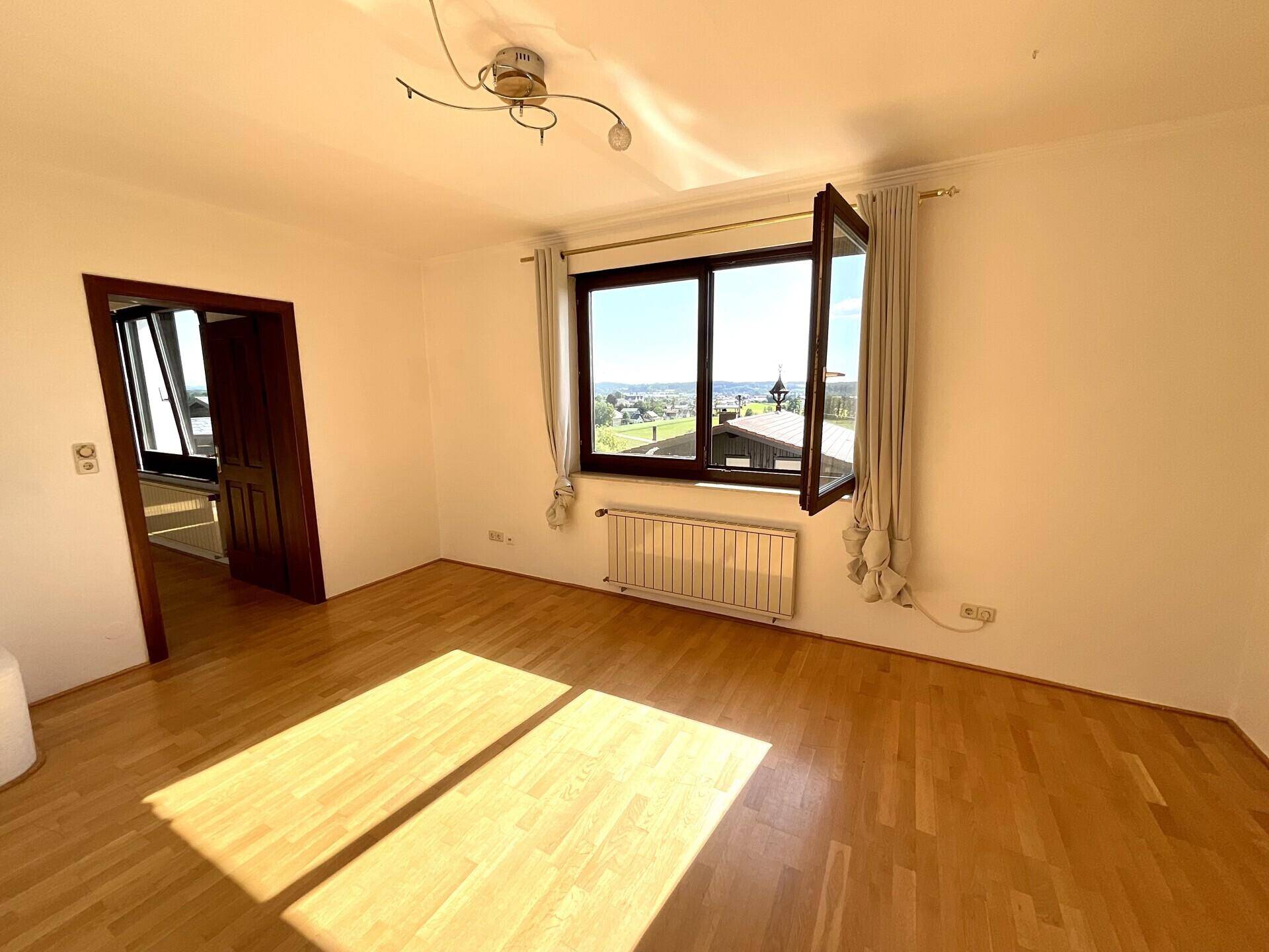 Helles Zimmer mit Parkettboden, großem Fenster und weitem Blick in die Landschaft.