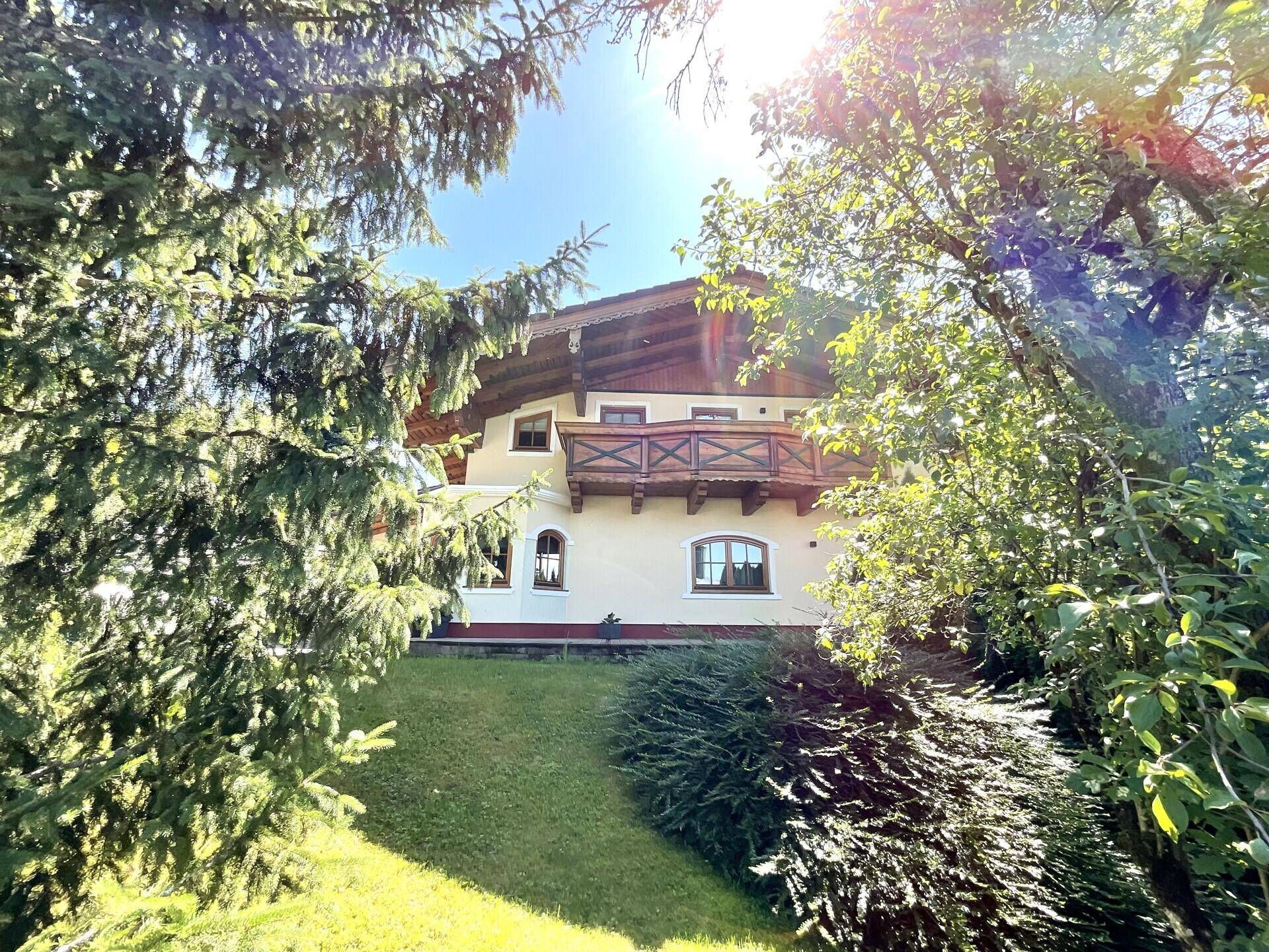 Traditionelles Haus mit Holzbalkon und üppiger grüner Umgebung unter strahlendem Sonnenschein.