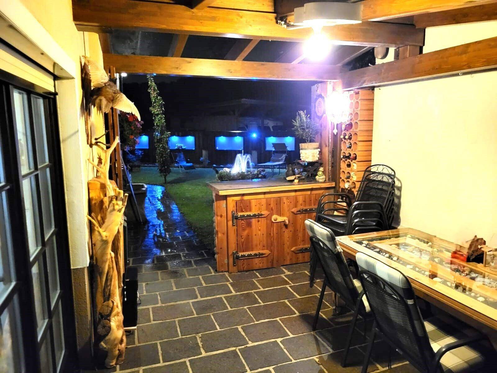 Überdachte Terrasse mit Sitzgelegenheiten und Blick auf den beleuchteten Garten mit Springbrunnen.