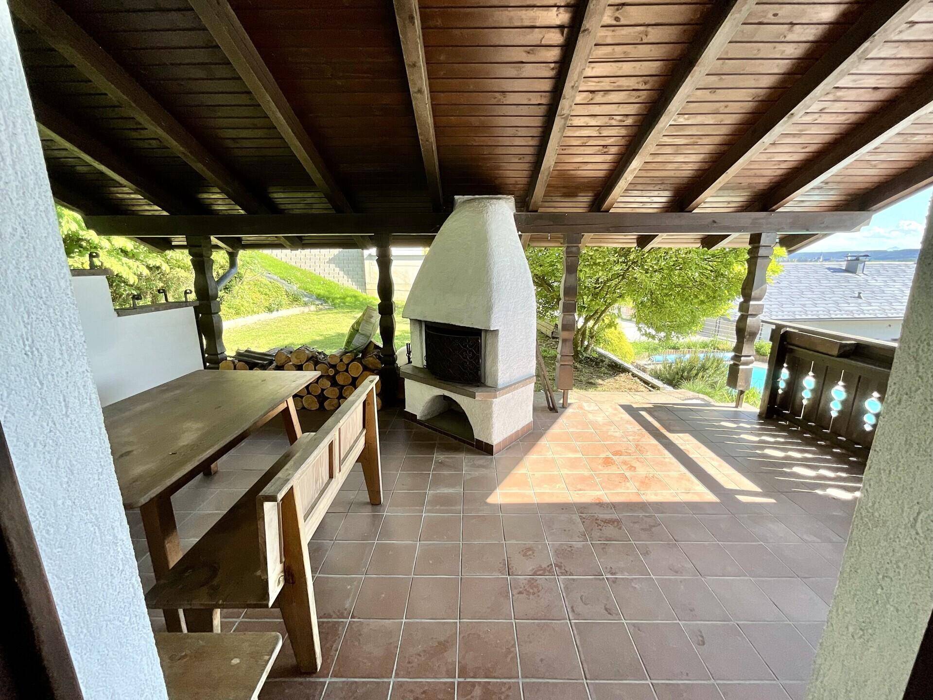 Überdachte Terrasse mit rustikalem Holztisch, Außenkamin und Blick ins Grüne.