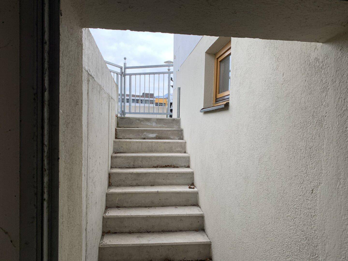 Betontreppe führt von einem Untergeschoss ins Freie, mit Metallgeländer und Fenster.