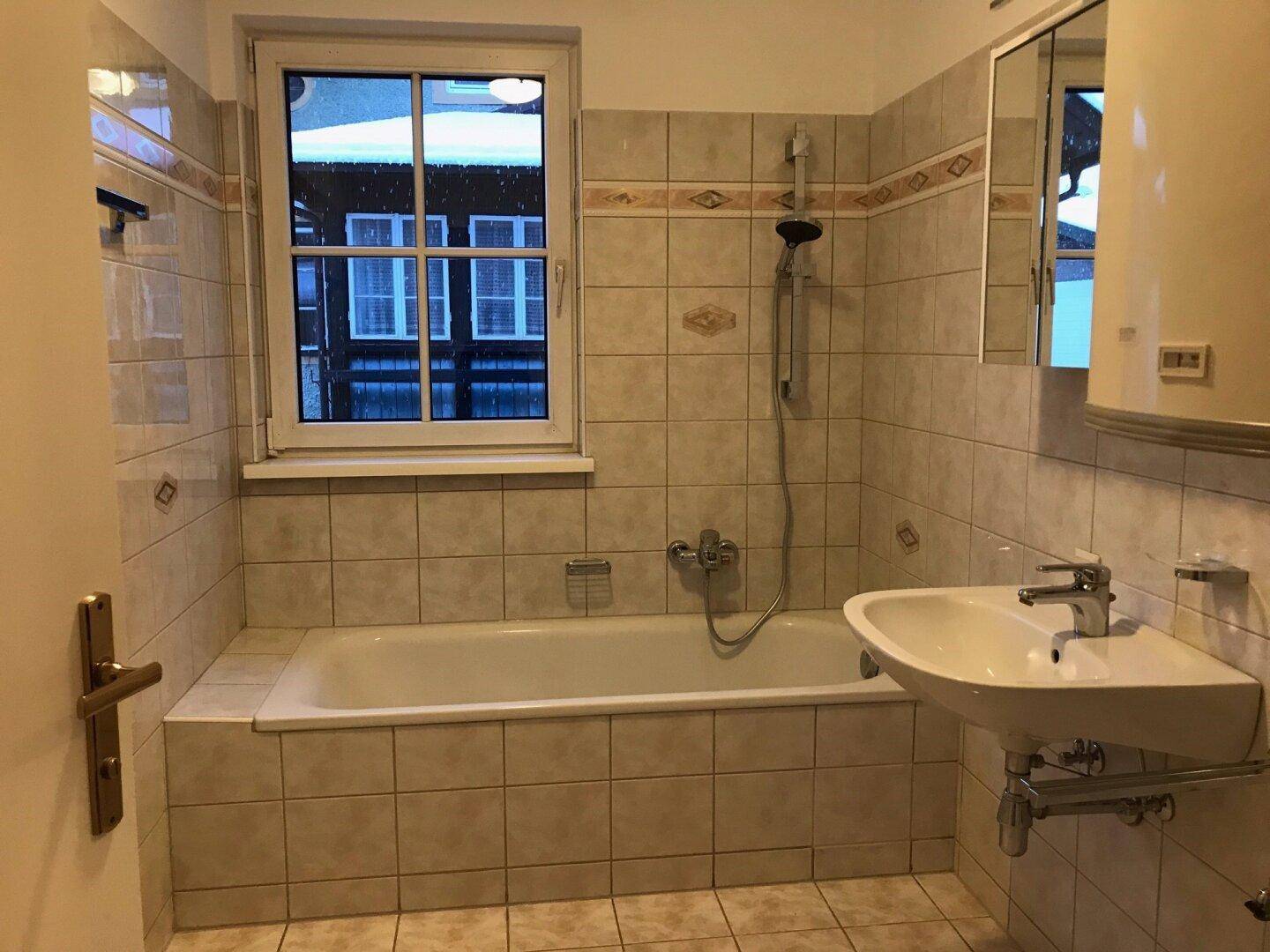 Gefliestes Badezimmer mit Badewanne, Duschfunktion, Waschbecken und Fenster.