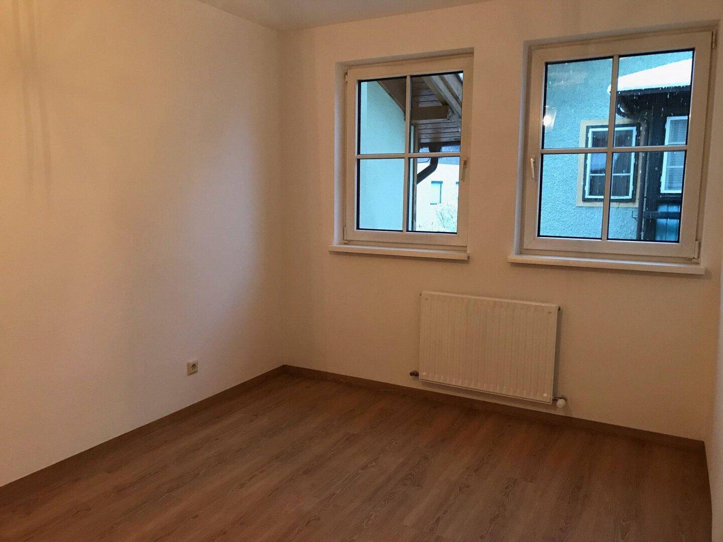 Leeres Zimmer mit Laminatboden, weißen Wänden und zwei Fenstern, ideal als Schlafzimmer.