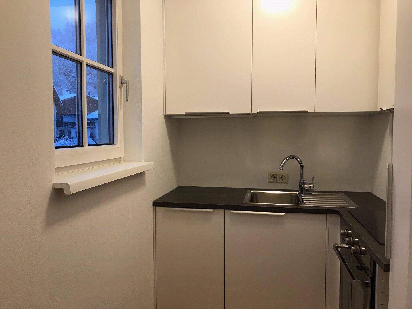 Kleine, moderne Einbauküche mit weißen Schränken, dunkler Arbeitsplatte und Fenster.