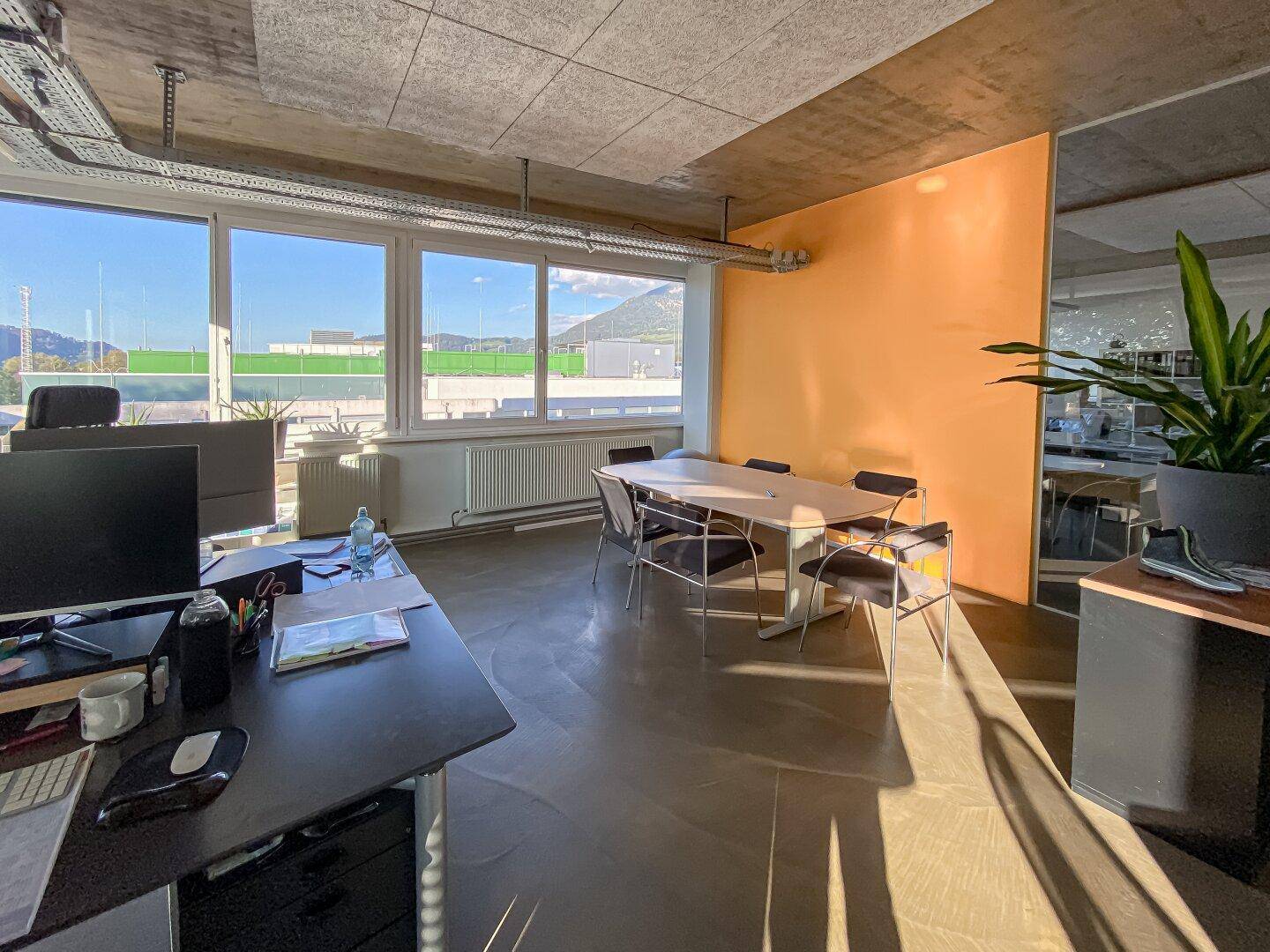 Bürobereich mit Besprechungstisch, farbiger Akzentwand und großen Fenstern mit Bergblick.