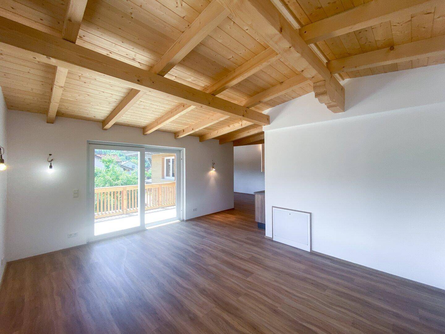 Wohnzimmer mit Zugang zum Balkon, Laminatboden und sichtbarer Holzdeckenkonstruktion.