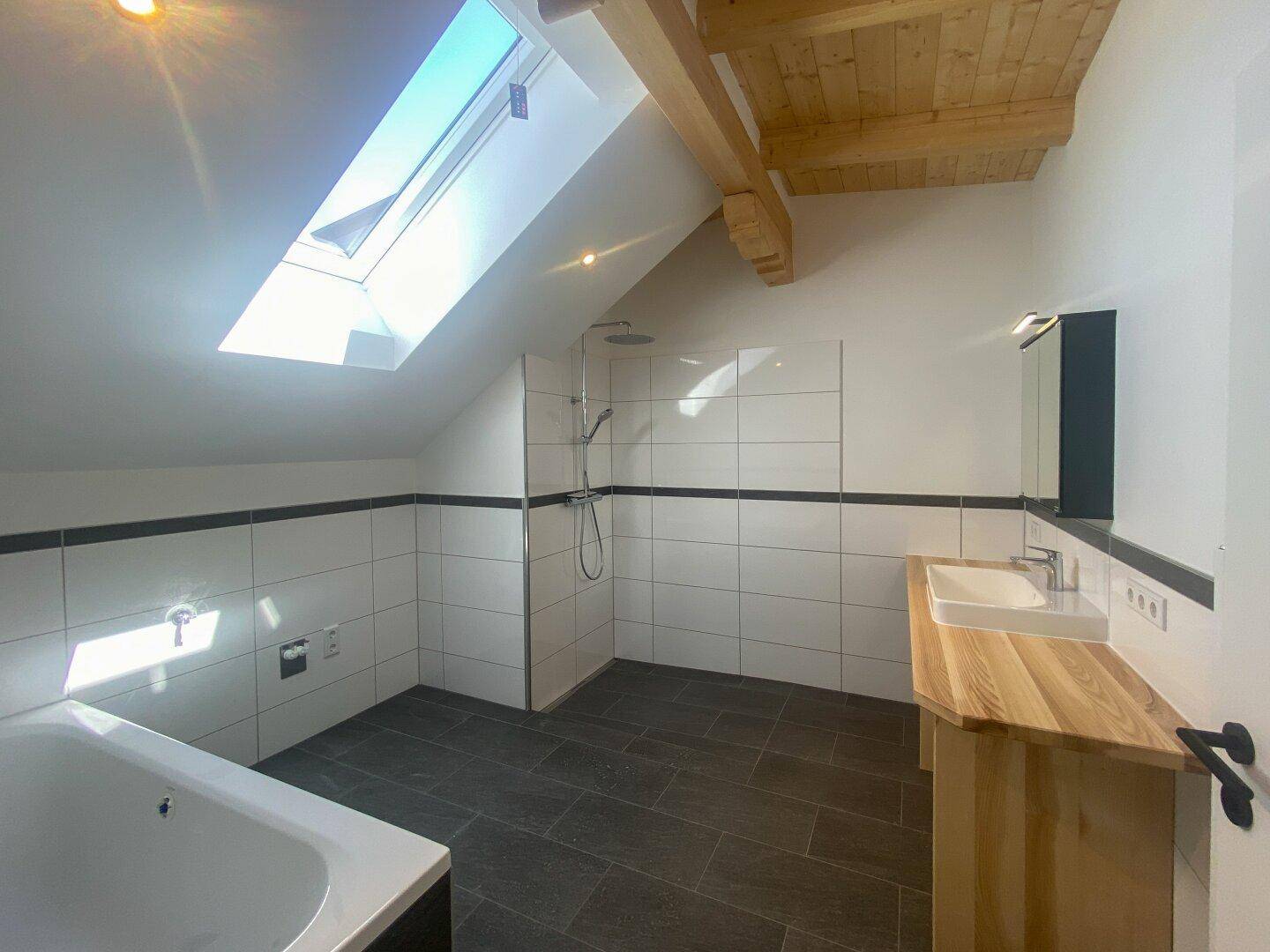 Helles Badezimmer mit Dachfenster, Dusche, Badewanne, Doppelwaschbecken und dunklen Bodenfliesen.