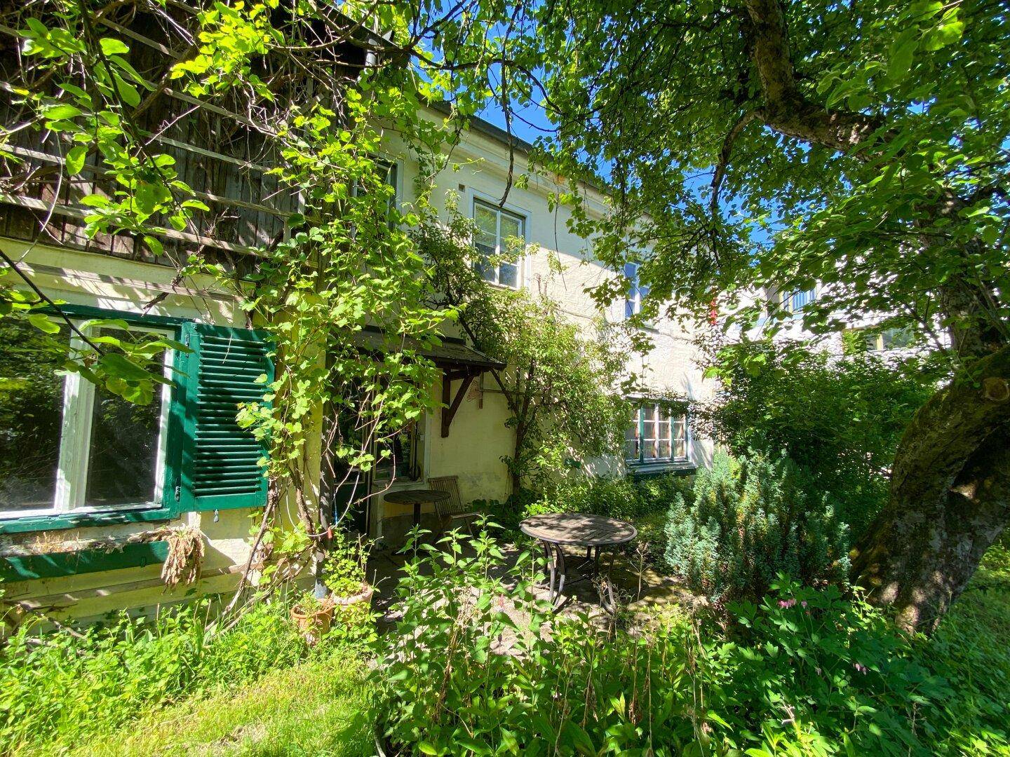Gemütlicher Sitzbereich im Freien unter Bäumen, umgeben von üppigem Grün am Haus.