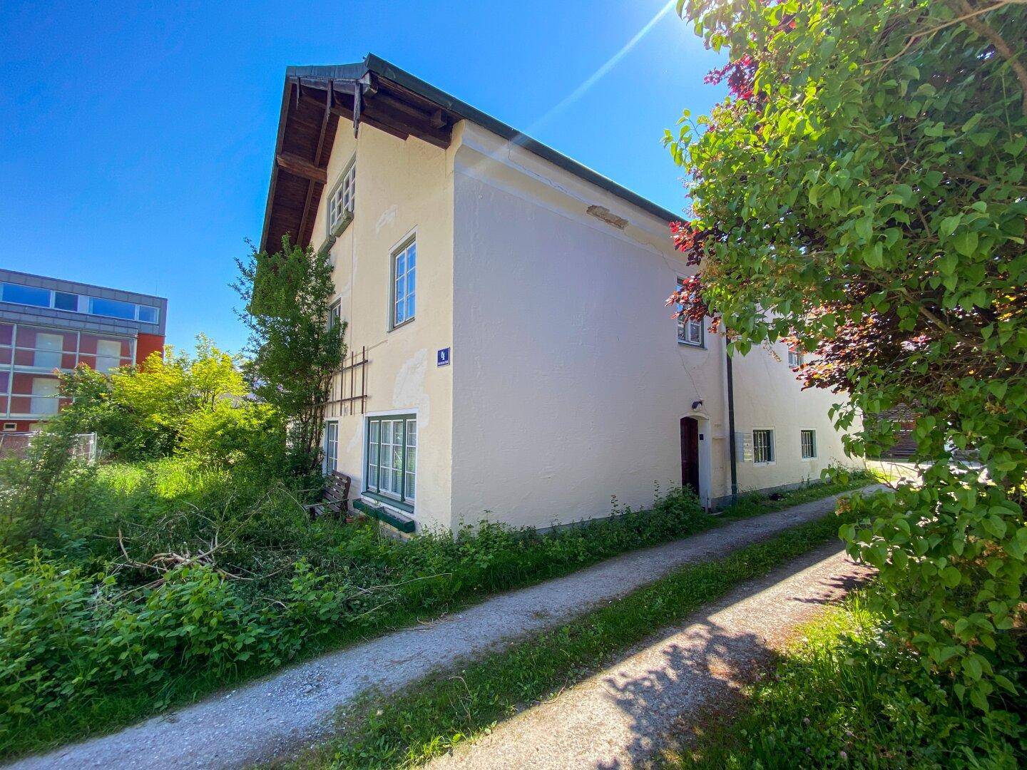 Seitenansicht des Hauses mit heller Fassade und Zufahrt, umgeben von Grünflächen.