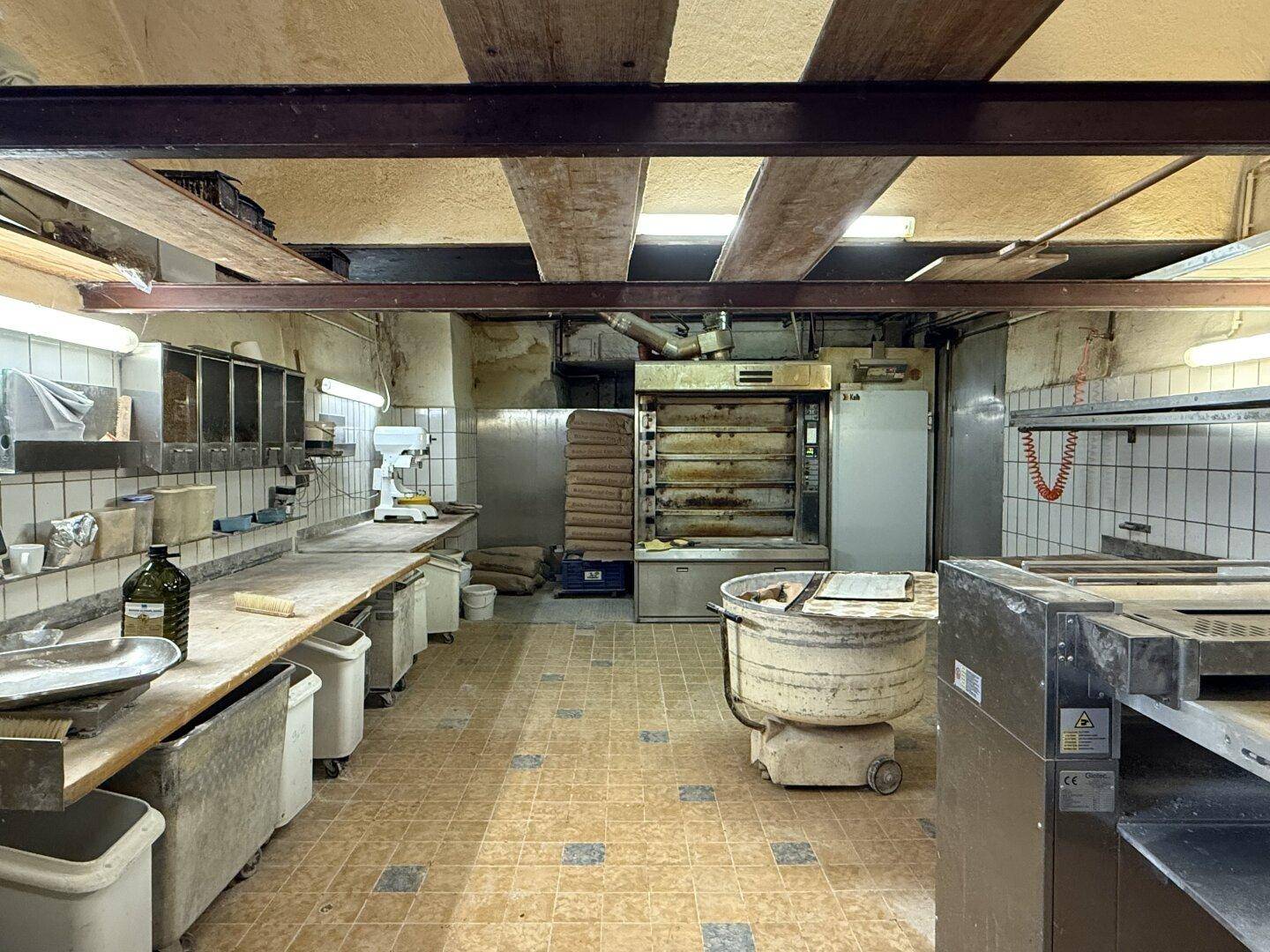 Große Bäckereiküche mit professionellen Öfen, Arbeitsflächen und Teigknetmaschinen unter Holzbalken.