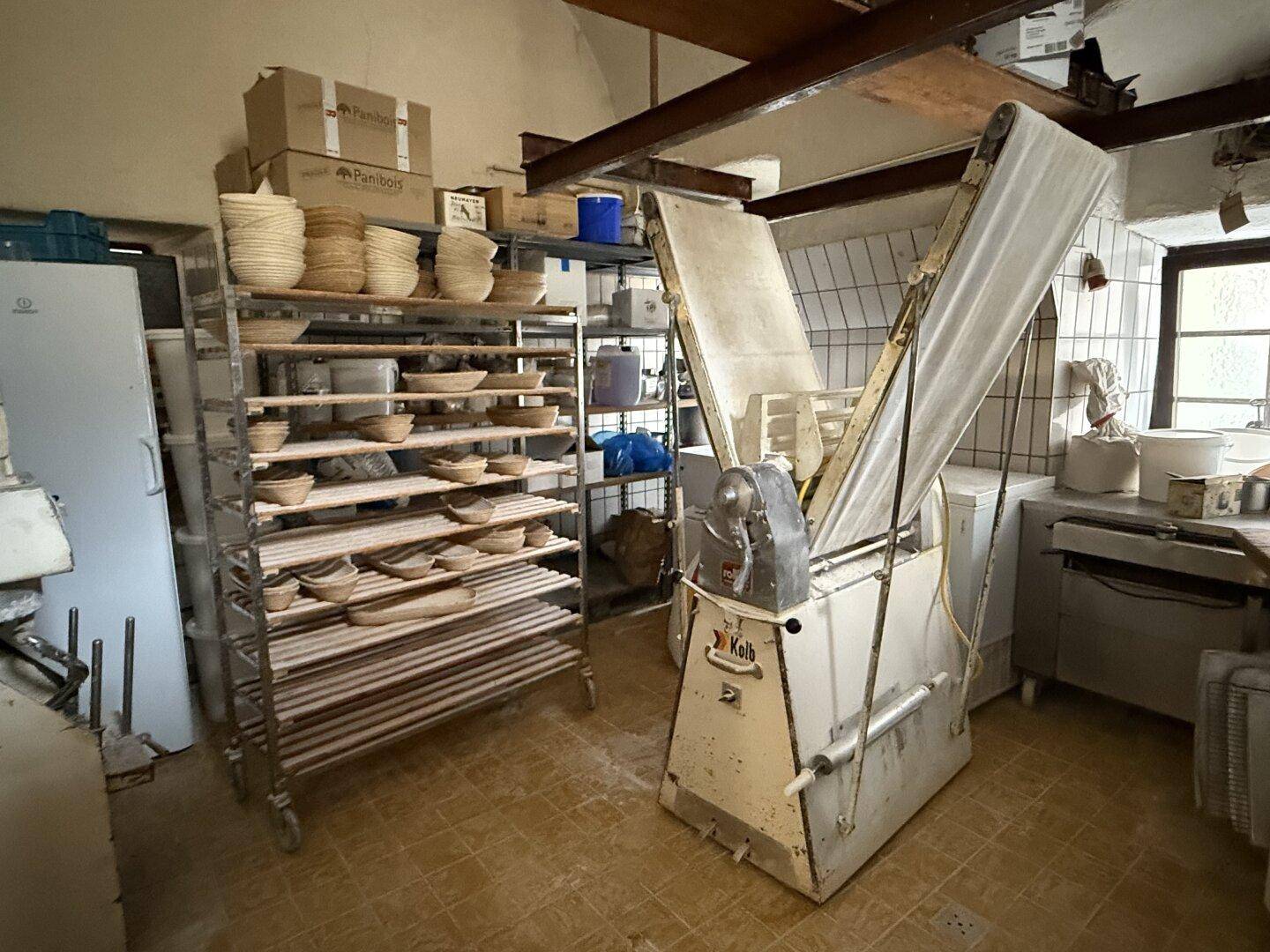 Bereich der Bäckerei mit Regalen voller Körbe und einer Teigausrollmaschine für die Produktion.