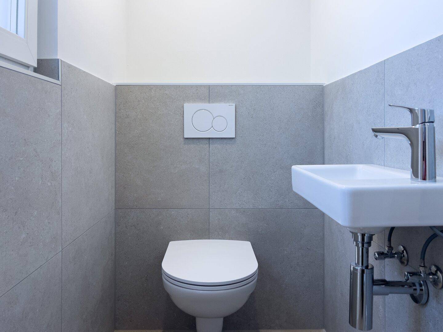 Modernes Gäste-WC mit grauen Fliesen, wandhängendem WC und kleinem Waschbecken.