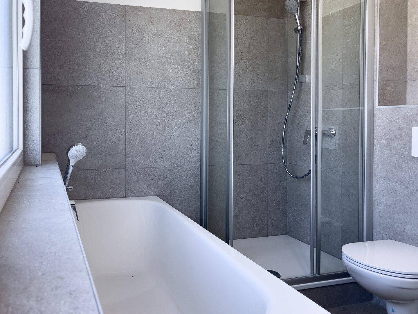 Modernes Badezimmer mit Badewanne, Dusche und grauen Fliesen an Wänden und Boden.