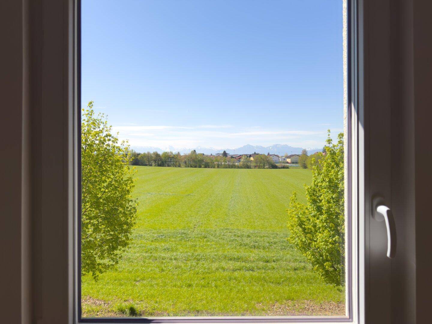 Weitblick aus dem Fenster über grüne Felder und ferne Berge unter blauem Himmel.