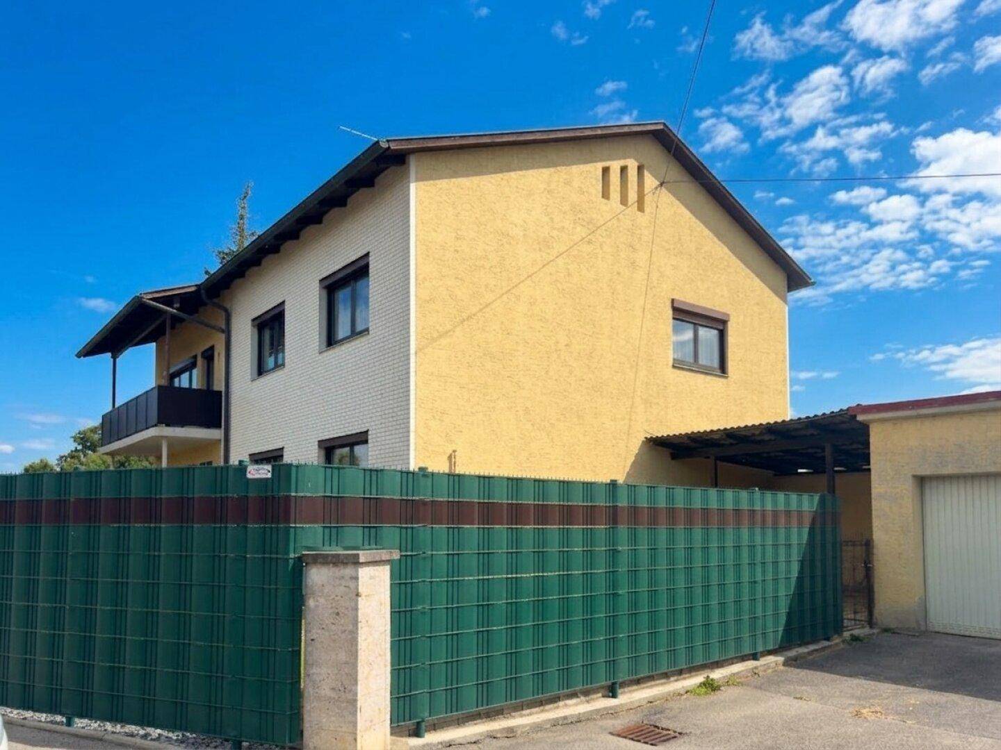 Seitenansicht des Hauses mit gelber Fassade und grünem Zaun, angrenzend an eine Garage.