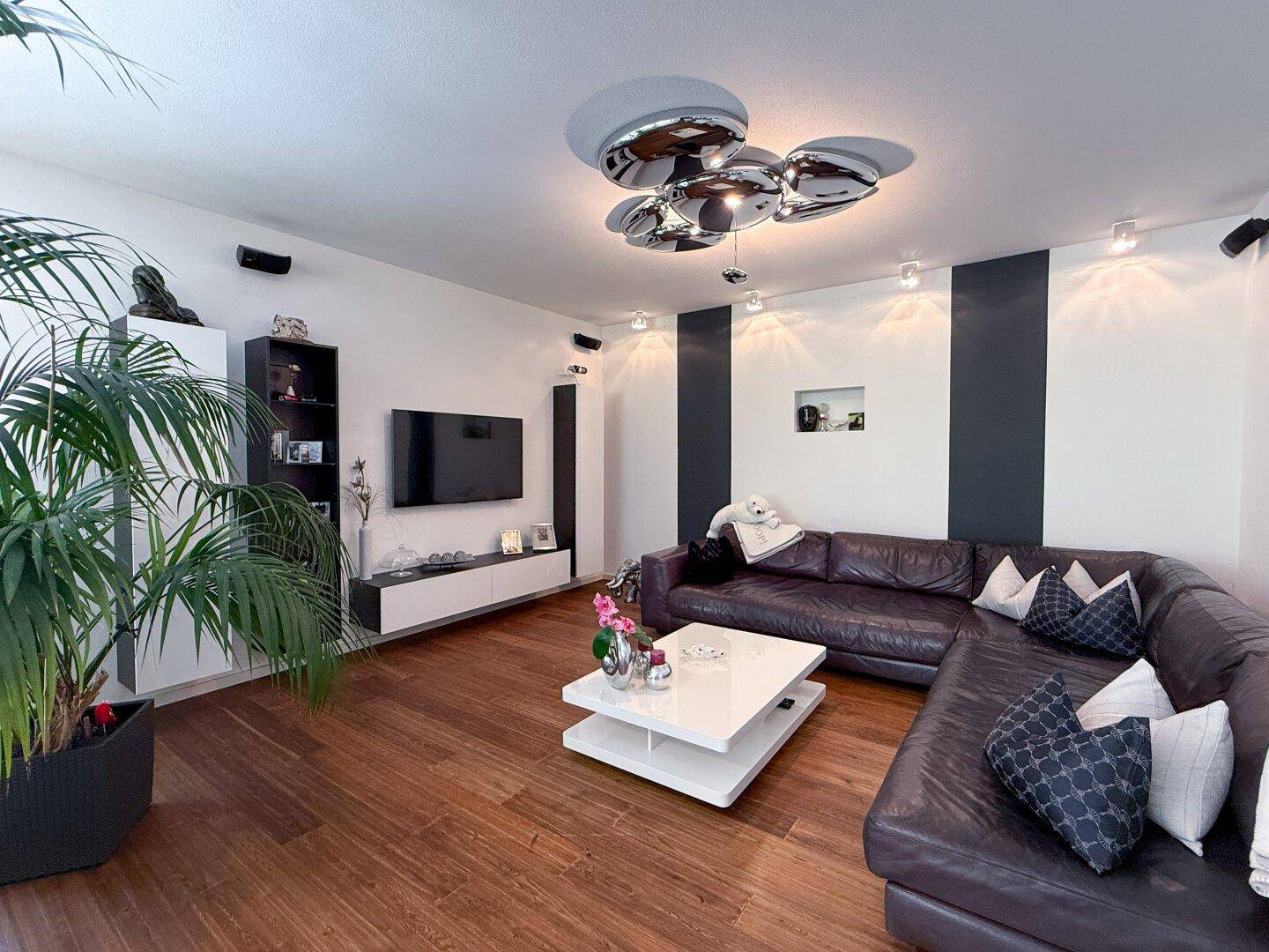 Gemütliches Wohnzimmer mit dunklem Ledersofa, modernem Couchtisch und Holzboden, dekoriert mit einer großen Palme.