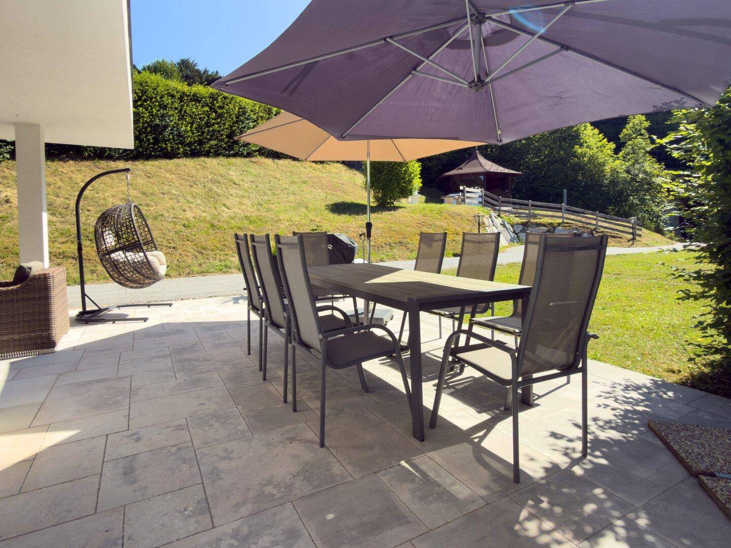 Große gepflasterte Terrasse mit Esstisch, Stühlen und Sonnenschirm, ideal für Mahlzeiten im Freien.