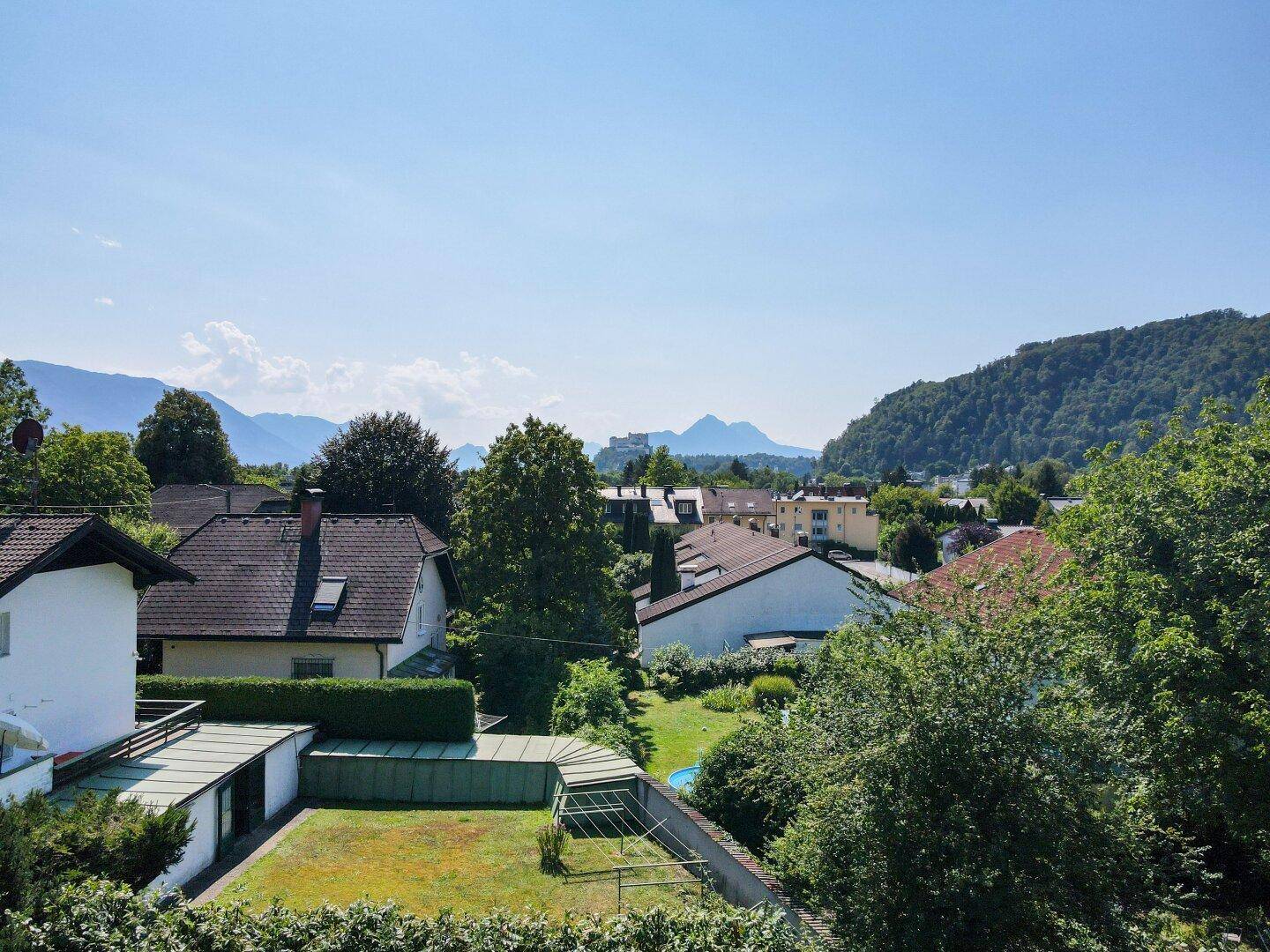 Weitläufiger Blick über Dächer, grüne Bäume und eine Berglandschaft mit einer Burg in der Ferne.