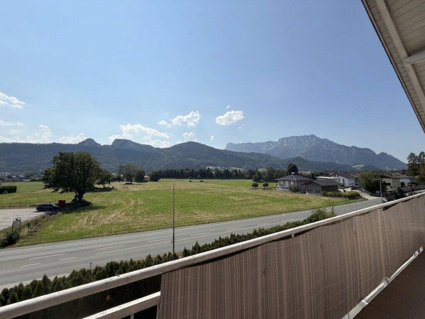 Weitläufiger Ausblick vom Balkon auf grüne Felder, eine Straße und majestätische Berge.