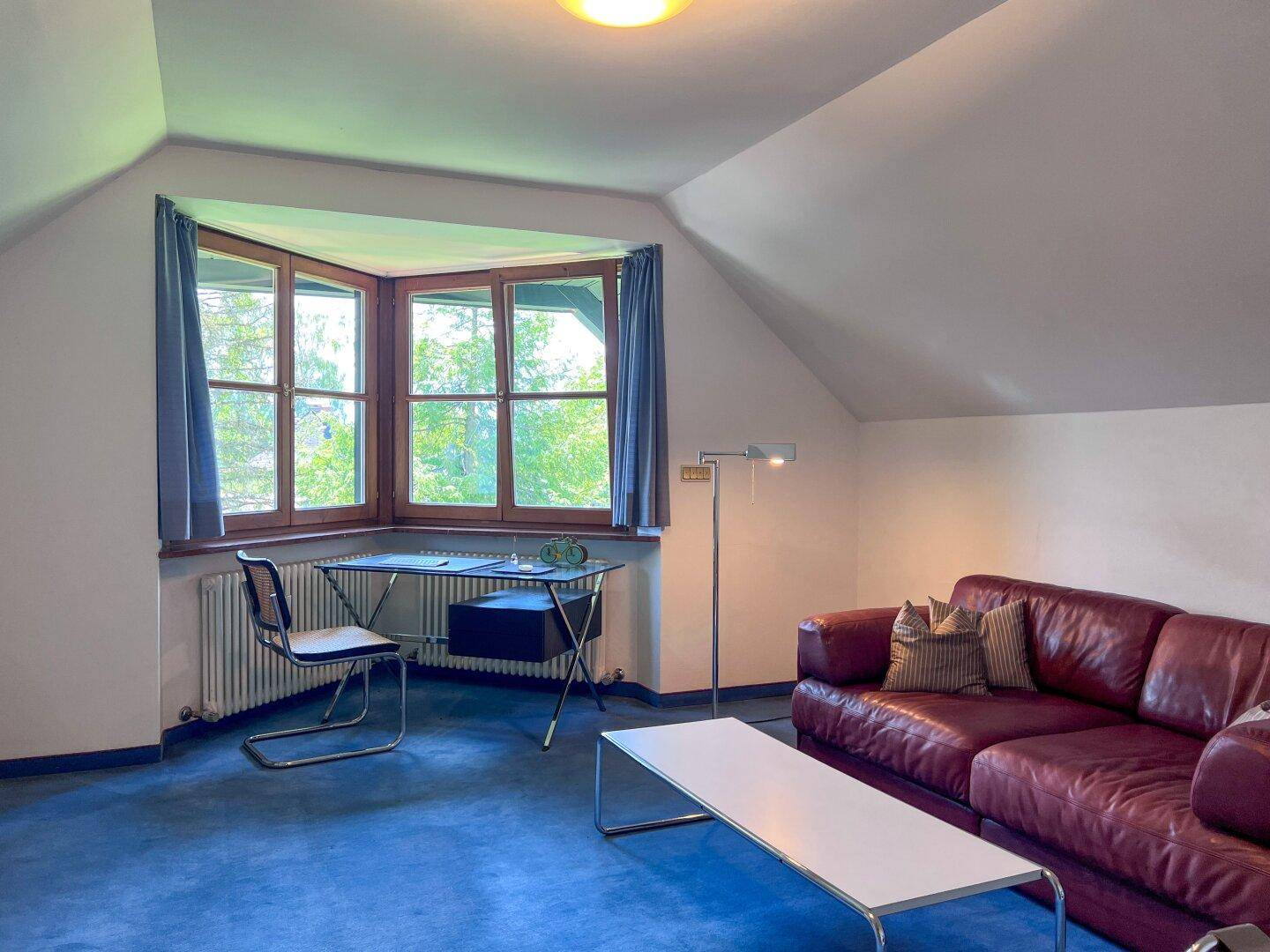 Arbeitszimmer oder Wohnbereich im Dachgeschoss mit blauem Teppich, Erkerfenster und Ledersofa.