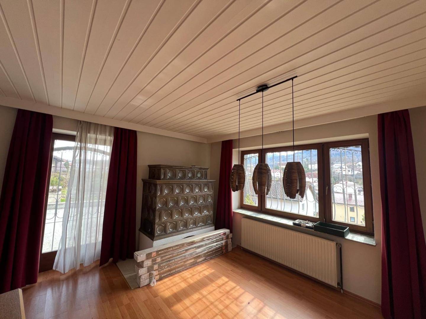 Gemütliches Zimmer mit traditionellem Kachelofen, Holzdecken und großen Fenstern mit rotem Vorhang.