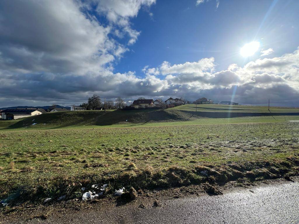 Weitläufige grüne Wiesen unter einem dramatischen Himmel mit Sonnenschein und entfernten Gebäuden.