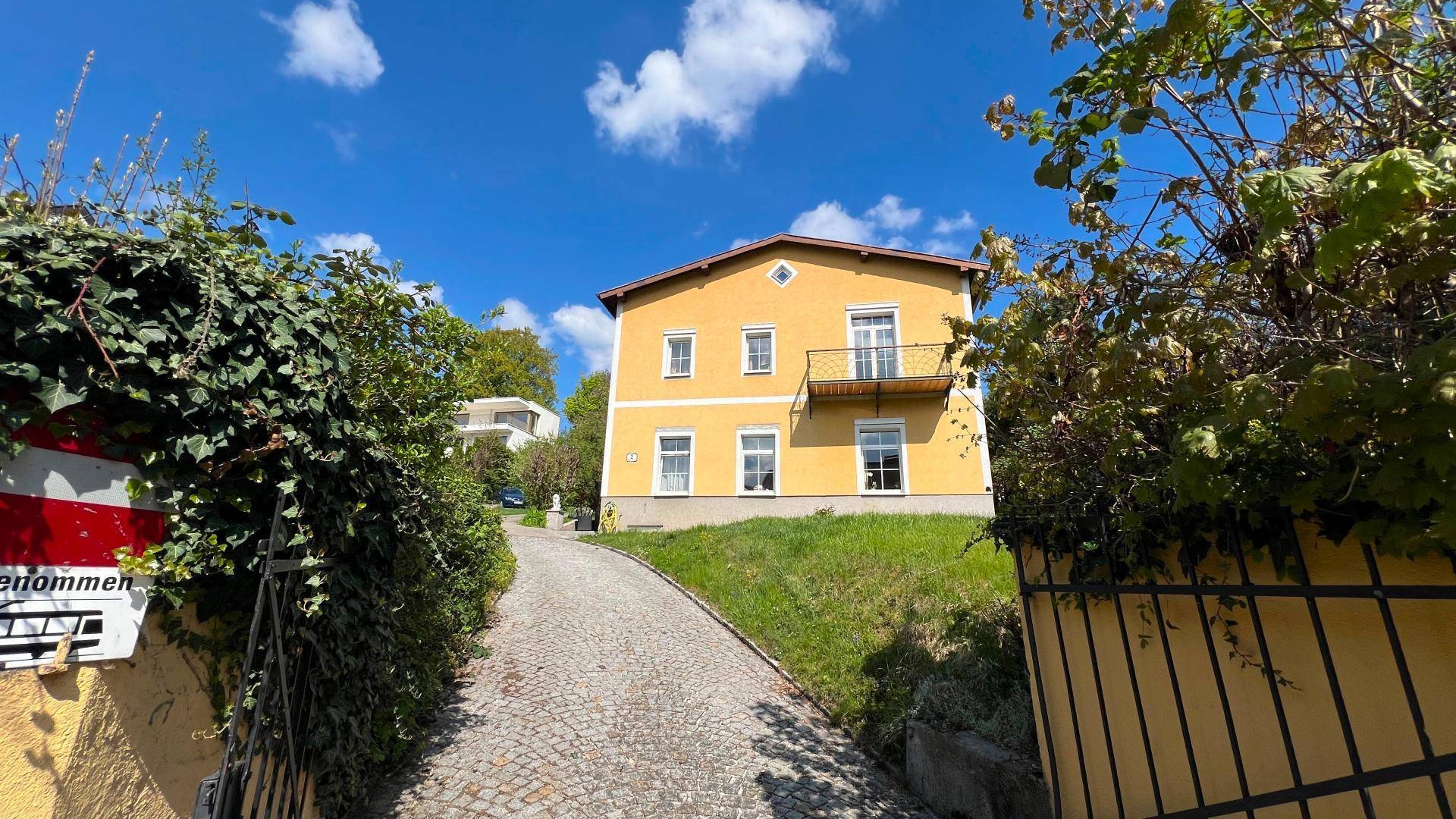 Gelbes Haus mit kleinem Balkon und Auffahrt, umgeben von grüner Vegetation unter blauem Himmel.