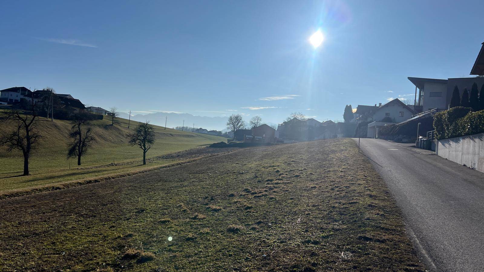 Sonnige Straße neben einem Feld mit Häusern und Bergen in der Ferne.