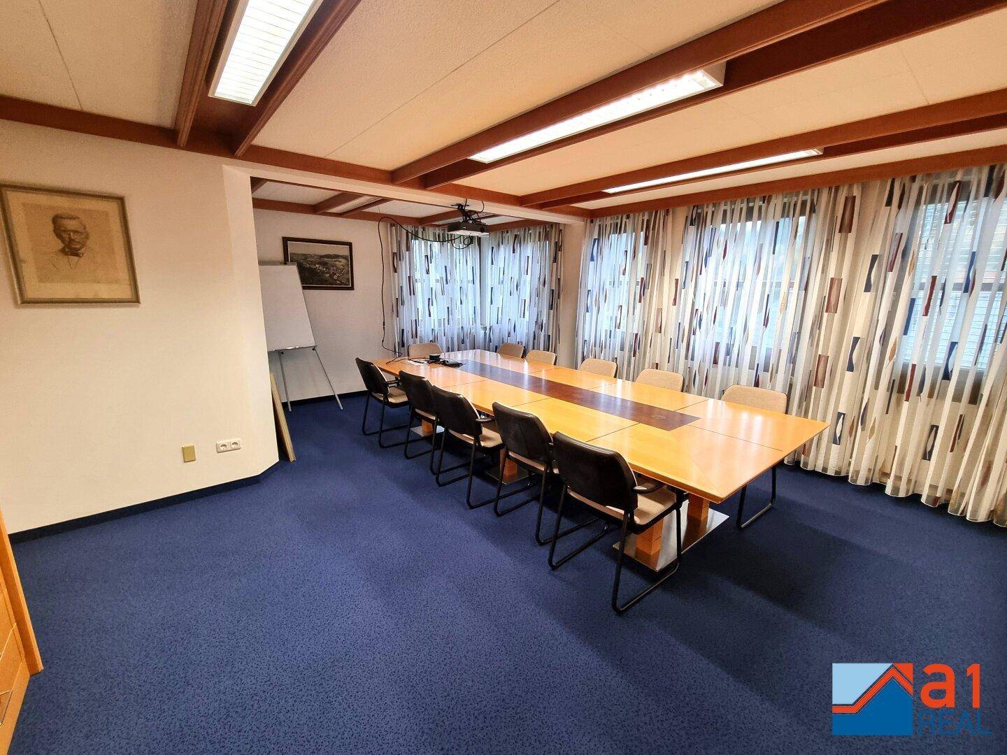 Weitläufiger Konferenzraum mit blauem Teppichboden, langem Holztisch und großen Fenstern für viel Tageslicht.