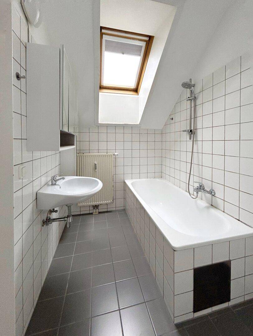 Badezimmer mit Badewanne, weißen Fliesen, grauen Bodenfliesen und Dachfenster.