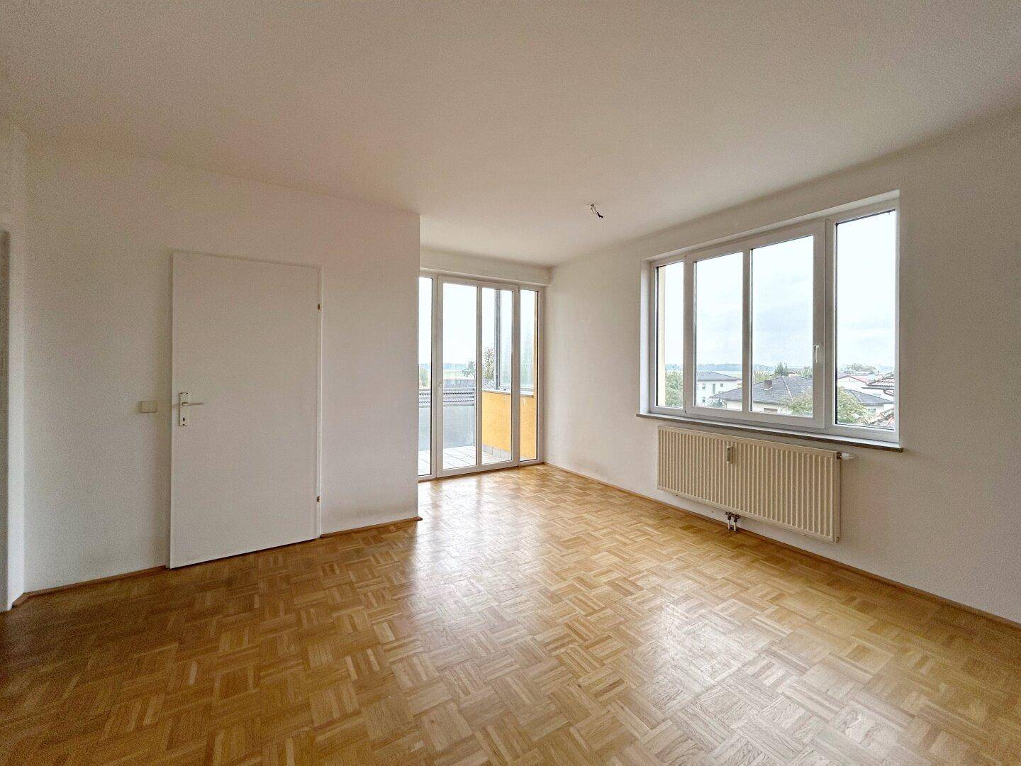 Helles Wohnzimmer mit Parkettboden, großen Fenstern und Zugang zu einem Balkon.