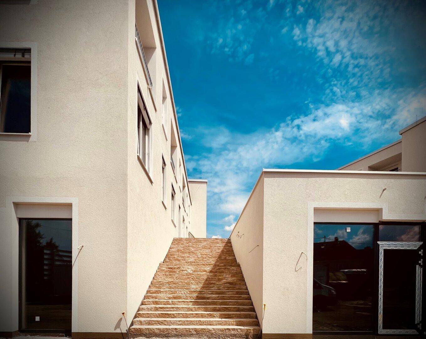 Moderne Gebäudefassade mit heller Putzoberfläche und einer Außentreppe unter blauem Himmel.
