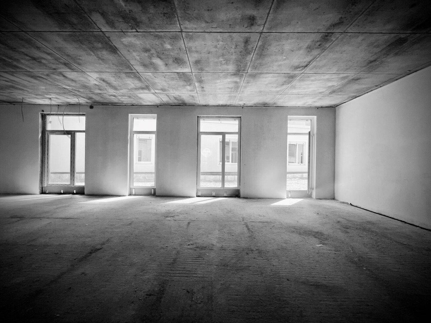 Rohbau eines hellen Gewerberaums in Schwarz-Weiß mit mehreren Fensteröffnungen und Betonboden.