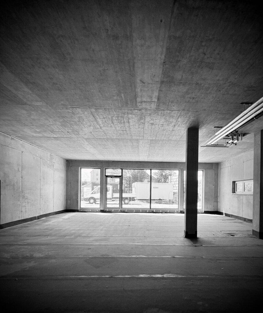 Unfertiger Gewerberaum in Schwarz-Weiß mit Sichtbetondecke und großen Fensterfronten zur Straße.