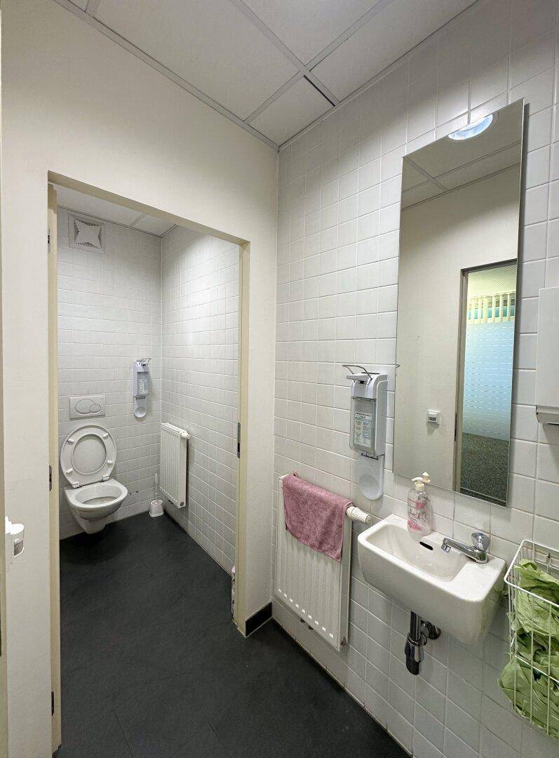 Einfache Toilette mit weißen Fliesen, Waschbecken und Wand-WC, ausgestattet mit Heizkörper.