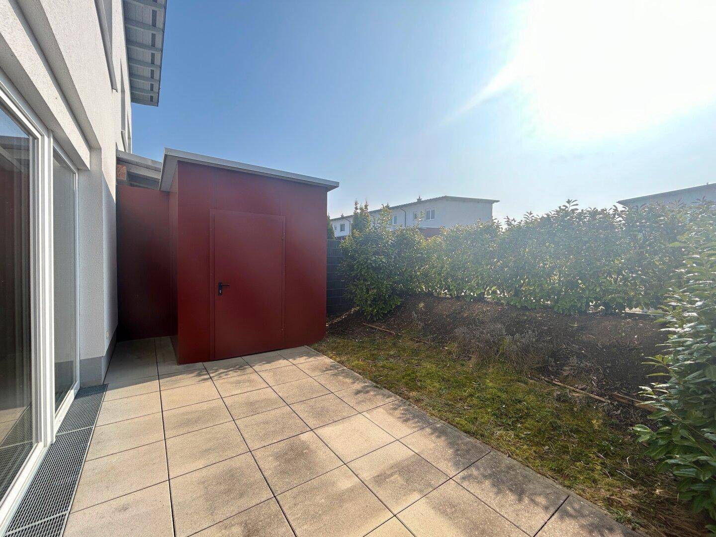 Sonniger Terrassenbereich mit Fliesenboden, Rasenfläche und einem praktischen Geräteschuppen.