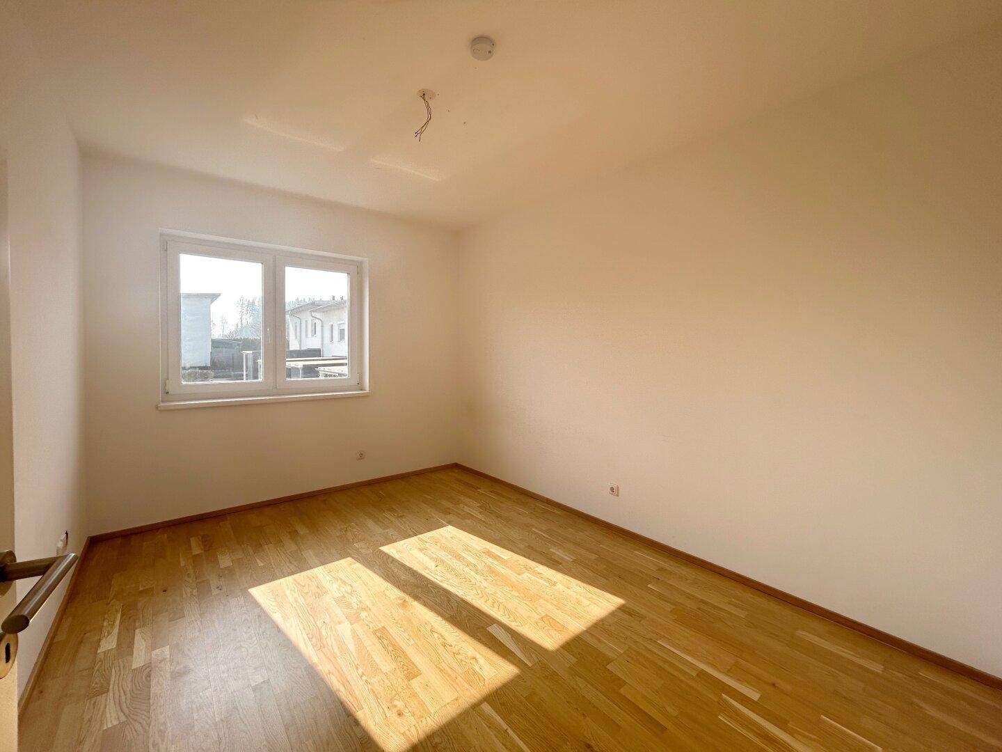 Helles Schlafzimmer mit Parkettboden und Fenster, das viel Tageslicht hereinlässt.