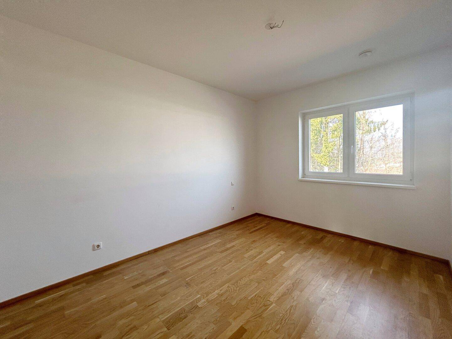 Geräumiges Schlafzimmer mit hellem Parkettboden und einem Fenster, das eine grüne Aussicht bietet.