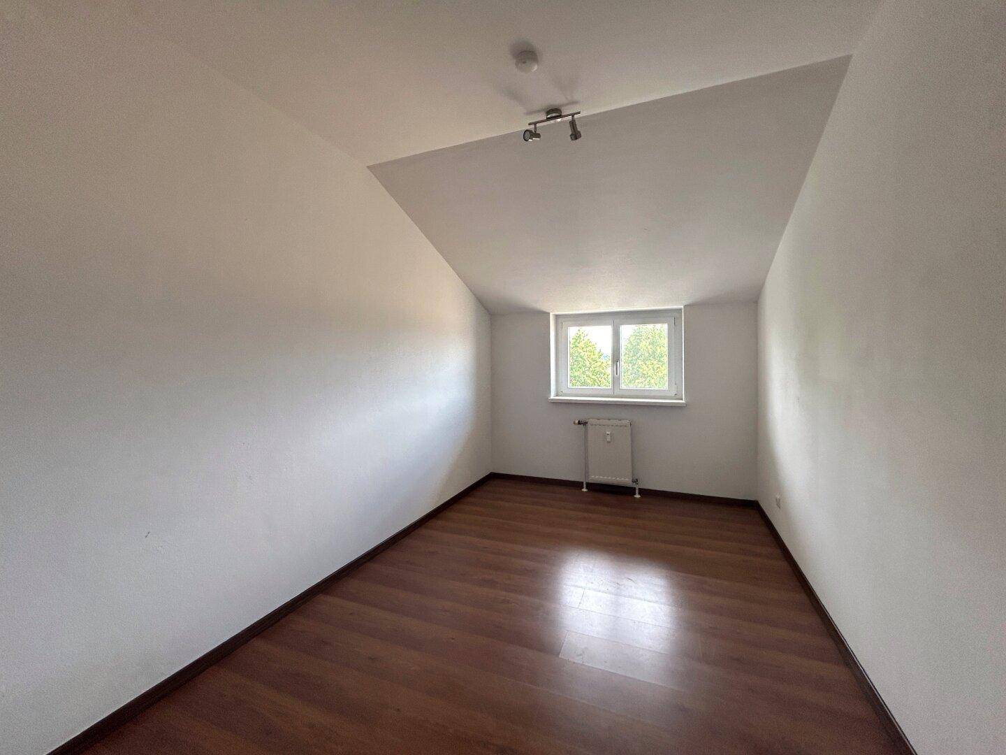 Helles Zimmer mit Laminatboden, Dachschräge und einem Fenster mit Blick ins Grüne.